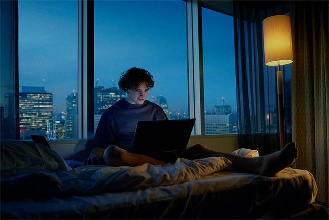 Kille sitter med laptop i sängen i svagt upplyst rum.